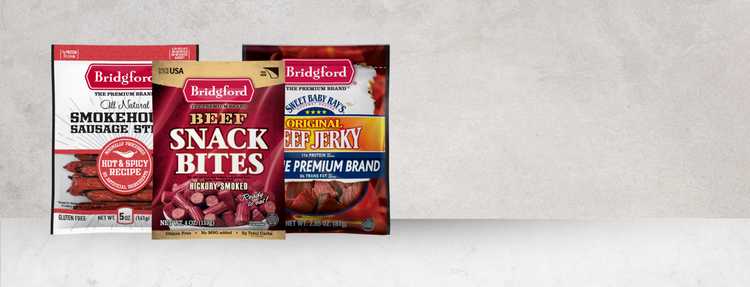 Three Bridgetown snack packages arranged side by side on a light gray textured background. The packages include Bridgeford Smokehouse Sausage Sticks, Bridgeford Snack Bites, and Bridgeford Smoked Beef Jerky.