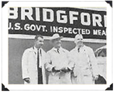 Vintage black and white photo of three men in white coats in front of a Bridgford U.S. Govt Inspected Meats sign.
