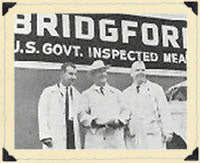 Black and white vintage photo of three inspectors standing in front of a Bridgford US Govt Inspected Meats sign.